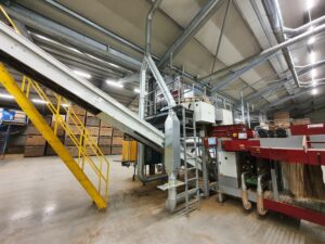 Produktionslinie mit Förderband und Auswurfsystem zur Kartoffelverarbeitung, mit Blick auf Rohre, Maschinen und Lagerkisten in der Halle.