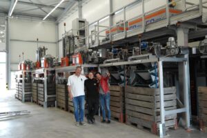 Trois employés de Gugger-Guillod posent devant la ligne d'emballage automatisée, avec des caisses en bois et des machines de tri.