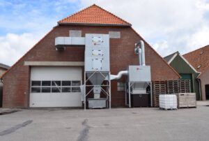 Jongejans Luchttechniek's extraction and filter system is installed against the outside wall of an agricultural shed, including air ducts and a collection unit.